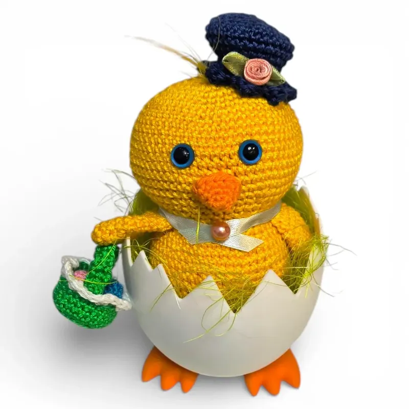 Poussin au crochet jaune avec chapeau haut-de-forme bleu et panier de Pâques dans son œuf sur pieds.